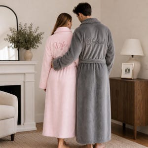 May include: A pair of plush, full-length robes featuring a textured collar and cuff design. The light pink robe is embroidered with the word Wife on the back, while the dark gray robe is embroidered with the word Husband on the back.