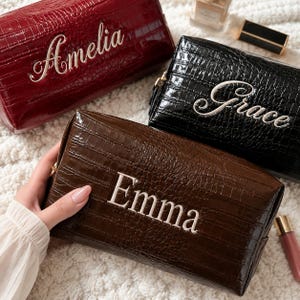 May include: Three rectangular cosmetic bags with a glossy crocodile-embossed texture. The bags are dark red, black, and dark brown. Each bag features a name embroidered in white script or block lettering: Amelia on the red, Grace on the black, and Emma on the brown bag. Each bag has a gold-toned zipper closure.