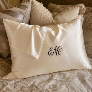 Op de afbeelding: Een rechthoekige, crèmekleurige kussensloop van zijde met een gladde, glanzende afwerking. De kussensloop is voorzien van een zwart geborduurd monogram met de letters EMC in een cursief lettertype, gecentreerd aan de rechteronderzijde.