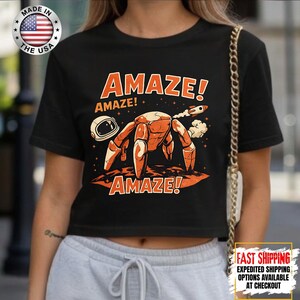May include: Black crop top with an orange and white graphic of a crab-like creature, an astronaut helmet, and a rocket ship. The word "AMAZE!" is printed in orange above and below the graphic. The shirt is made in the USA.
