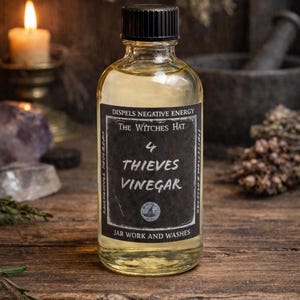 May include: A clear glass bottle with a black cap, filled with a yellow liquid. The label reads "4 Thieves Vinegar" and "Dispel Negative Energy". The bottle sits on a wooden surface, with a lit candle and other items in the background.