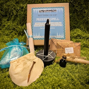 May include: A collection of items for a "Freeze Spell" kit, including a black candle in a pentagram holder, a scroll, a small bottle, a nail, and a small bag. The kit is packaged in a brown box with text that reads "The Witches Hat".