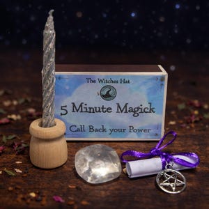Op de afbeelding: Een zilverkleurige, gedraaide kaars in een houten houder, een heldere kristal, een rol met een pentagrambedel en een doos met de tekst "5 Minute Magick". De items zijn gerangschikt op een houten oppervlak.