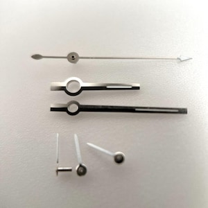 Puede incluir: Un conjunto de agujas de reloj plateadas, que incluyen una larga y delgada con punta de flecha, y varias agujas más cortas con extremos circulares y puntiagudos. Para la reparación o personalización de relojes.