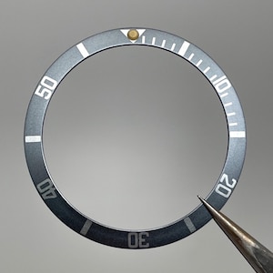 May include: A dark gray watch bezel with white markings for minutes and numerals at 10-minute intervals. The bezel has a gold-colored dot at the top and is being held by a silver tool.