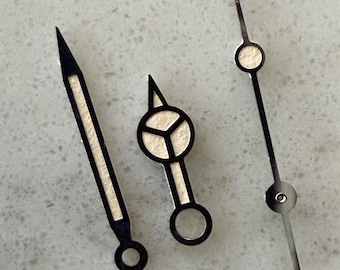 May include: A set of three watch hands featuring a long pointed hour hand, a shorter Mercedes-style hand with a circular center, and a thin, straight second hand. Each hand is constructed from dark metal with off-white luminous inserts.