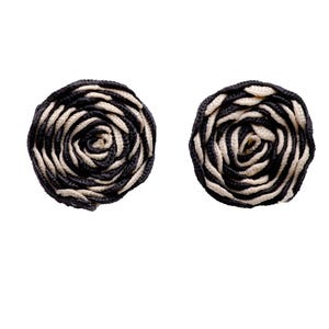 Puede incluir: Dos pendientes circulares hechos a mano con un diseño floral. Cada pendiente presenta un patrón en espiral de material tejido negro y crema, que se asemeja a una rosa. Los pendientes están sobre un fondo blanco liso.