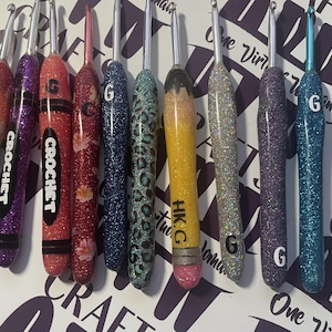 May include: A collection of nine metal crochet hooks with custom glitter-coated handles. The handles feature various designs including a rainbow gradient with text reading GODS PROMISE, floral patterns, leopard print, and a yellow pencil motif.