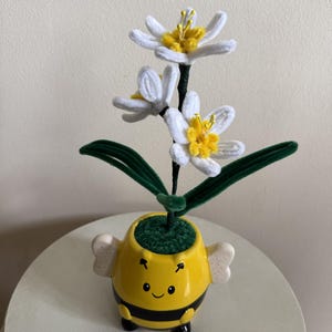Puede incluir: Arreglo floral artesanal con narcisos blancos y amarillos. Las flores están colocadas en una maceta de cerámica amarilla con forma de abeja, con rayas negras y una cara sonriente. El arreglo incluye hojas verdes y un tallo oscuro.