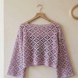 May include: A light purple, open-weave crochet top with long sleeves. The top is hanging on a wooden hanger. The crochet pattern features a floral design, creating an airy and delicate look. The sleeves are wide and the neckline is a boat neck.