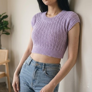 May include: A lavender crochet crop top with a ribbed hem and neckline. The short-sleeved top has an open, textured stitch pattern. The top is paired with blue jeans.
