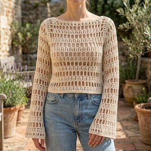 May include: A beige long-sleeved crochet top featuring a wide boat neckline and an open, grid-like stitch pattern. The garment has a cropped length and flared sleeves, paired with light blue denim jeans.