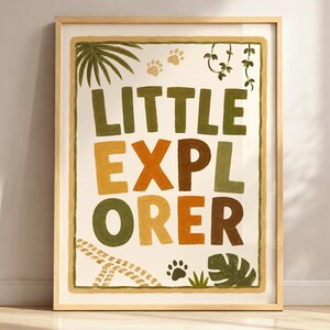 Könnte beinhalten: Ein gerahmter Kunstdruck mit dem Schriftzug "LITTLE EXPLORER" in einer verspielten, farbenfrohen Schriftart. Das Design umfasst Dschungel-Illustrationen wie Pfotenabdrücke, Blätter und Ranken, vor einem cremefarbenen Hintergrund. Der Rahmen ist hellbraun.