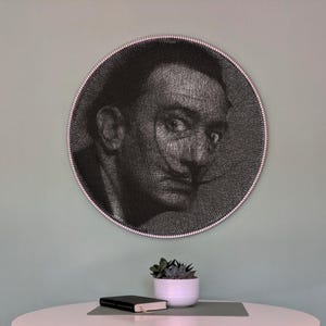 Könnte beinhalten: Ein schwarz-weißes String-Art-Porträt von Salvador Dalí in einem kreisförmigen Rahmen mit einem rosa und weißen Rand. Das Kunstwerk hängt an einer hellgrünen Wand. Eine kleine Sukkulentenpflanze in einem weißen Topf und ein schwarzes Notizbuch stehen auf einem weißen Tisch.