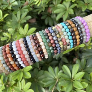 May include: A collection of beaded bracelets in various colors, including orange, black, white, pink, blue, and green. The bracelets are displayed on a tan cylindrical holder against a backdrop of green foliage. Each bracelet appears to be made of small, round beads.