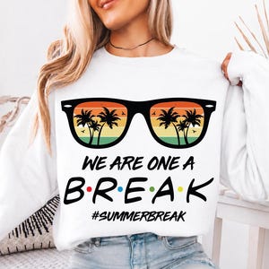 May include: White sweatshirt with a graphic of sunglasses with palm trees and a sunset design. The text reads "WE ARE ONE A BREAK" with the word "BREAK" in colorful letters. Below the text is the hashtag #SUMMERBREAK.