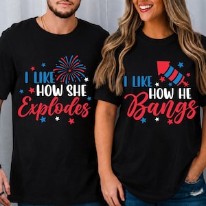 May include: Two black short-sleeved t-shirts featuring red, white, and blue graphics. One shirt displays a firework and the text I LIKE HOW SHE EXPLODES. The other shirt displays a rocket and the text I LIKE HOW HE BANGS, both with star accents.