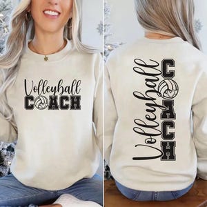 May include: A cream-coloured crewneck sweatshirt featuring black text and graphics. The front displays the words Volleyball Coach with a volleyball icon. The back features the same text and a volleyball icon arranged in a vertical layout.
