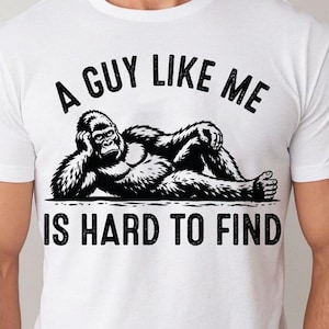 Pode incluir: Uma t-shirt branca de manga curta com um gráfico preto de um gorila deitado de lado. Acima do gorila, o texto diz A GUY LIKE ME, e abaixo do gráfico, o texto diz IS HARD TO FIND numa fonte preta, em negrito e desgastada.