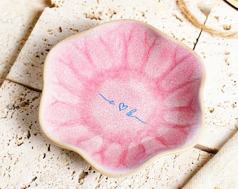 May include: A small, scalloped-edge ceramic trinket dish with a glossy pink crackle glaze finish. The centre of the dish features a delicate blue line drawing of the letters e and k connected by a small heart symbol.