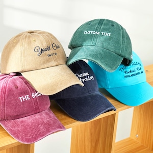 May include: A collection of five baseball caps in various colours including tan, dark green, magenta, navy blue, and bright blue. Each cap features embroidered text such as Yacht Club, Custom Text, Custom Embroidery, and The William Cocktail Club.