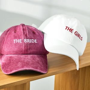 May include: A maroon baseball cap with the white embroidered text THE BRIDE and a white baseball cap with the red embroidered text THE GIRLS. Both caps feature a curved brim, a six-panel construction, and eyelets for ventilation on the crown.