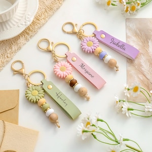 May include: Three personalised keychains featuring a gold-tone metal lobster clasp and ring, a silicone daisy flower charm, a series of wooden and silicone beads, and a faux leather strap with the names Harley, Morgane, and Isabella printed.