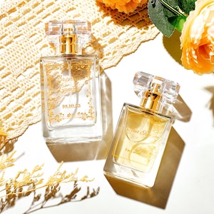 Op de afbeelding: Twee rechthoekige glazen parfumflesjes met doorzichtige doppen en gouden verstuivers. Het linkerflesje heeft een gouden bloemenkader met de tekst "Brenna Camp" en "10.10.25". Het rechterflesje toont de tekst "Aria Skye" en "I'm Loved".