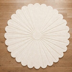 Puede incluir: Una alfombra circular de color blanco roto con forma de flor y numerosos p&eacute;talos redondeados que irradian desde un n&uacute;cleo central. La alfombra presenta una superficie texturizada con l&iacute;neas hundidas que definen cada p&eacute;talo en el dise&ntilde;o.