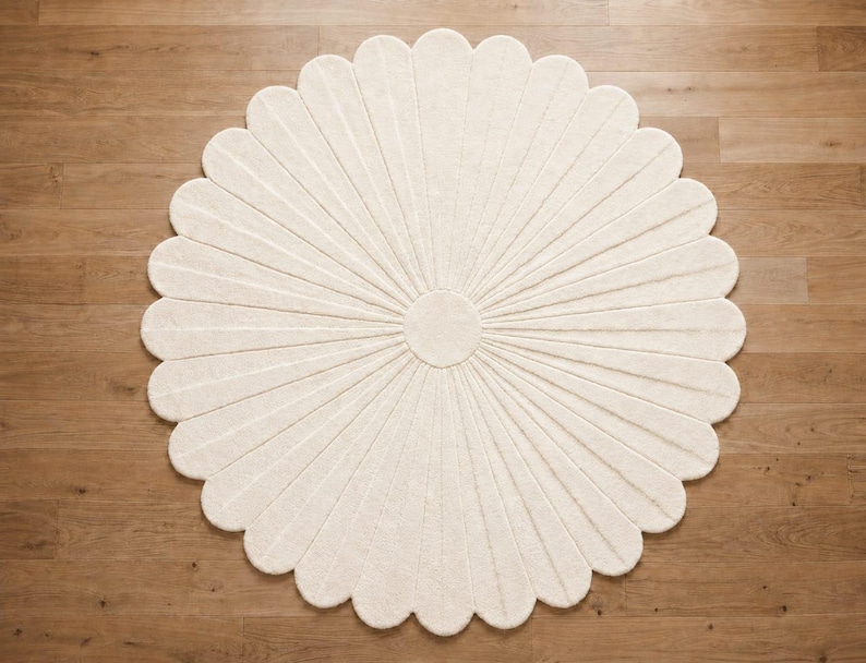 Puede incluir: Una alfombra circular de color blanco roto con forma de flor y numerosos p&eacute;talos redondeados que irradian desde un n&uacute;cleo central. La alfombra presenta una superficie texturizada con l&iacute;neas hundidas que definen cada p&eacute;talo en el dise&ntilde;o.