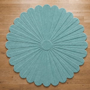 Puede incluir: Una alfombra circular de color verde azulado claro, con un patr&oacute;n radial de segmentos alargados en forma de p&eacute;talos que se extienden desde un n&uacute;cleo central. La alfombra tiene una superficie texturizada y un borde festoneado en forma de flor.