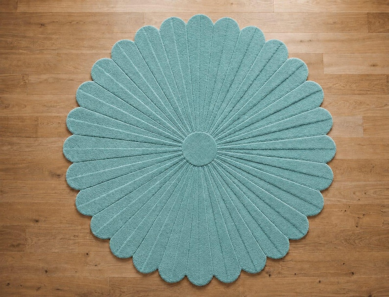 Puede incluir: Una alfombra circular de color verde azulado claro, con un patr&oacute;n radial de segmentos alargados en forma de p&eacute;talos que se extienden desde un n&uacute;cleo central. La alfombra tiene una superficie texturizada y un borde festoneado en forma de flor.