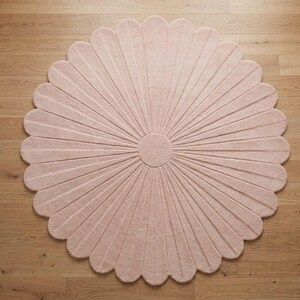 A pale pink round tufted rug shaped like a daisy with scalloped petals lies on a wooden floor.