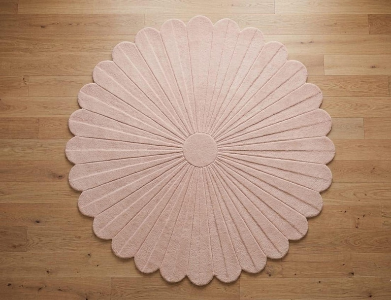 A pale pink round tufted rug shaped like a daisy with scalloped petals lies on a wooden floor.