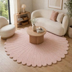 A large, circular, light pink area rug featuring a scalloped edge design that resembles flower petals. The rug has a textured, tufted surface with radiating lines extending from the centre towards the outer scalloped border.