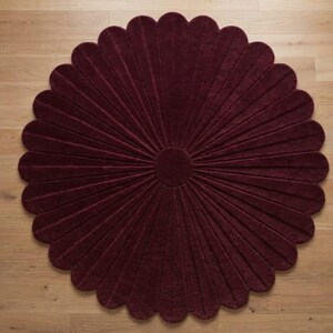 Puede incluir: Una alfombra circular de color burdeos intenso, con un patr&oacute;n radial de segmentos alargados en forma de p&eacute;talos que se extienden desde un centro circular s&oacute;lido. La alfombra tiene una superficie texturizada y un borde festoneado.