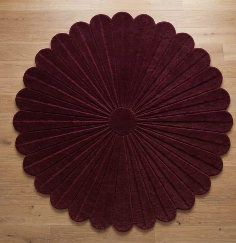 Puede incluir: Una alfombra circular de color burdeos intenso, con un patr&oacute;n radial de segmentos alargados en forma de p&eacute;talos que se extienden desde un centro circular s&oacute;lido. La alfombra tiene una superficie texturizada y un borde festoneado.