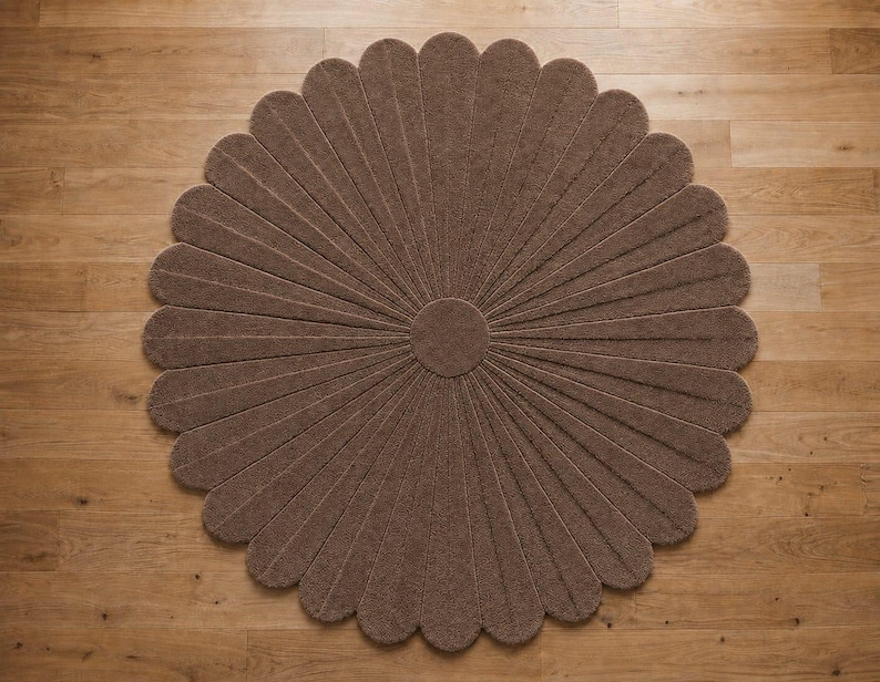 Puede incluir: Una alfombra circular marr&oacute;n con forma de flor, compuesta por numerosos p&eacute;talos alargados y redondeados que irradian desde un centro circular s&oacute;lido. La alfombra presenta una superficie texturizada y copetuda, dispuesta en un dise&ntilde;o floral.