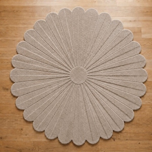Puede incluir: Una alfombra circular de color beige con un dise&ntilde;o de p&eacute;talos radiales texturizados que se asemeja a una flor grande. La alfombra tiene un n&uacute;cleo central circular con segmentos largos y estrechos que se extienden hacia afuera para crear un borde festoneado, proporcionando un revestimiento de suelo uniforme.