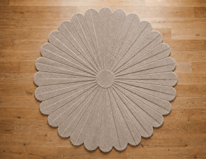 Puede incluir: Una alfombra circular de color beige con un dise&ntilde;o de p&eacute;talos radiales texturizados que se asemeja a una flor grande. La alfombra tiene un n&uacute;cleo central circular con segmentos largos y estrechos que se extienden hacia afuera para crear un borde festoneado, proporcionando un revestimiento de suelo uniforme.