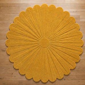 Puede incluir: Una alfombra circular de color amarillo mostaza con forma de flor, que presenta un disco central redondo y numerosos p&eacute;talos radiantes. La alfombra tiene una superficie texturizada y con mechones, con surcos profundos entre cada p&eacute;talo.