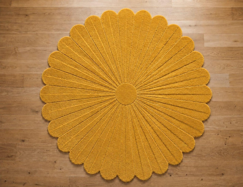 Puede incluir: Una alfombra circular de color amarillo mostaza con forma de flor, que presenta un disco central redondo y numerosos p&eacute;talos radiantes. La alfombra tiene una superficie texturizada y con mechones, con surcos profundos entre cada p&eacute;talo.