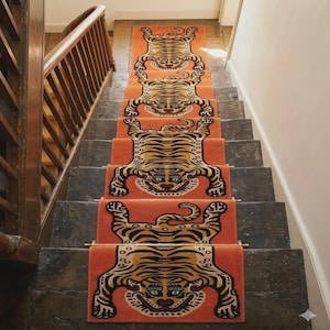 Puede incluir: Una alfombra para escaleras con un patrón repetitivo de tigres estilizados en negro y beige sobre un fondo naranja. La alfombra está fijada a los escalones de madera oscura con varillas metálicas en cada peldaño, creando una secuencia decorativa.