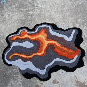 May include: An irregularly shaped area rug featuring a layered, topographic design. The rug displays concentric, flowing bands of color in shades of black, dark gray, light gray, and vibrant orange, creating a high-contrast, abstract pattern.