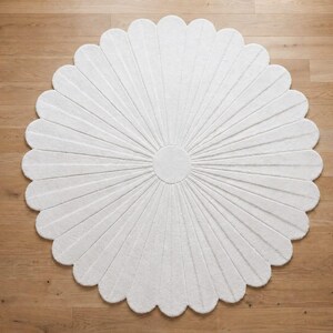 Puede incluir: Una alfombra circular blanca con forma de flor y numerosos p&eacute;talos redondeados que irradian desde un c&iacute;rculo central s&oacute;lido. La alfombra presenta una superficie texturizada con finas l&iacute;neas hundidas que se extienden desde el centro hasta el borde.