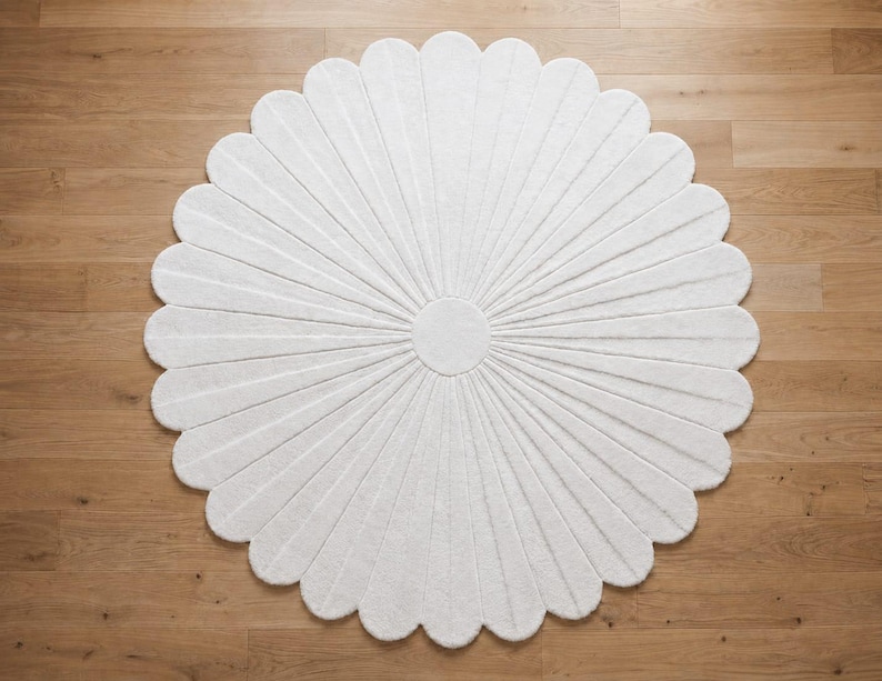 Puede incluir: Una alfombra circular blanca con forma de flor y numerosos p&eacute;talos redondeados que irradian desde un c&iacute;rculo central s&oacute;lido. La alfombra presenta una superficie texturizada con finas l&iacute;neas hundidas que se extienden desde el centro hasta el borde.