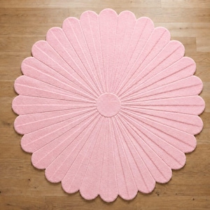 Puede incluir: Una alfombra circular de color rosa claro con forma de flor, compuesta por numerosos p&eacute;talos redondeados que irradian desde un n&uacute;cleo central. La alfombra presenta una superficie texturizada y acolchada con l&iacute;neas que separan los p&eacute;talos.