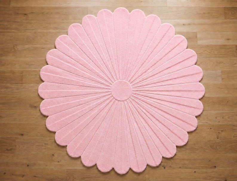 Puede incluir: Una alfombra circular de color rosa claro con forma de flor, compuesta por numerosos p&eacute;talos redondeados que irradian desde un n&uacute;cleo central. La alfombra presenta una superficie texturizada y acolchada con l&iacute;neas que separan los p&eacute;talos.