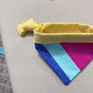 Può includere: Una bandana triangolare per animali domestici con un bordo superiore a pois gialli e bianchi e un nodo. Il corpo principale è composto da tre pannelli di tessuto diagonali in blu reale con un piccolo motivo floreale, azzurro a pois bianchi e magenta.