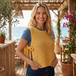May include: A mustard yellow sleeveless crochet vest featuring a square-patterned knit design with side ties at the waist. The vest is worn over a plain blue short-sleeved t-shirt and paired with dark blue denim jeans, creating a layered look.